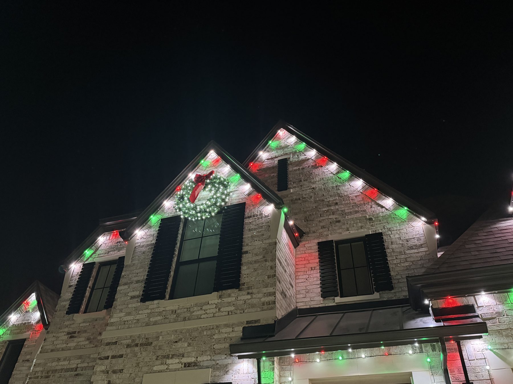 Two-story home in red, green and white holiday lighting with a lit wreath