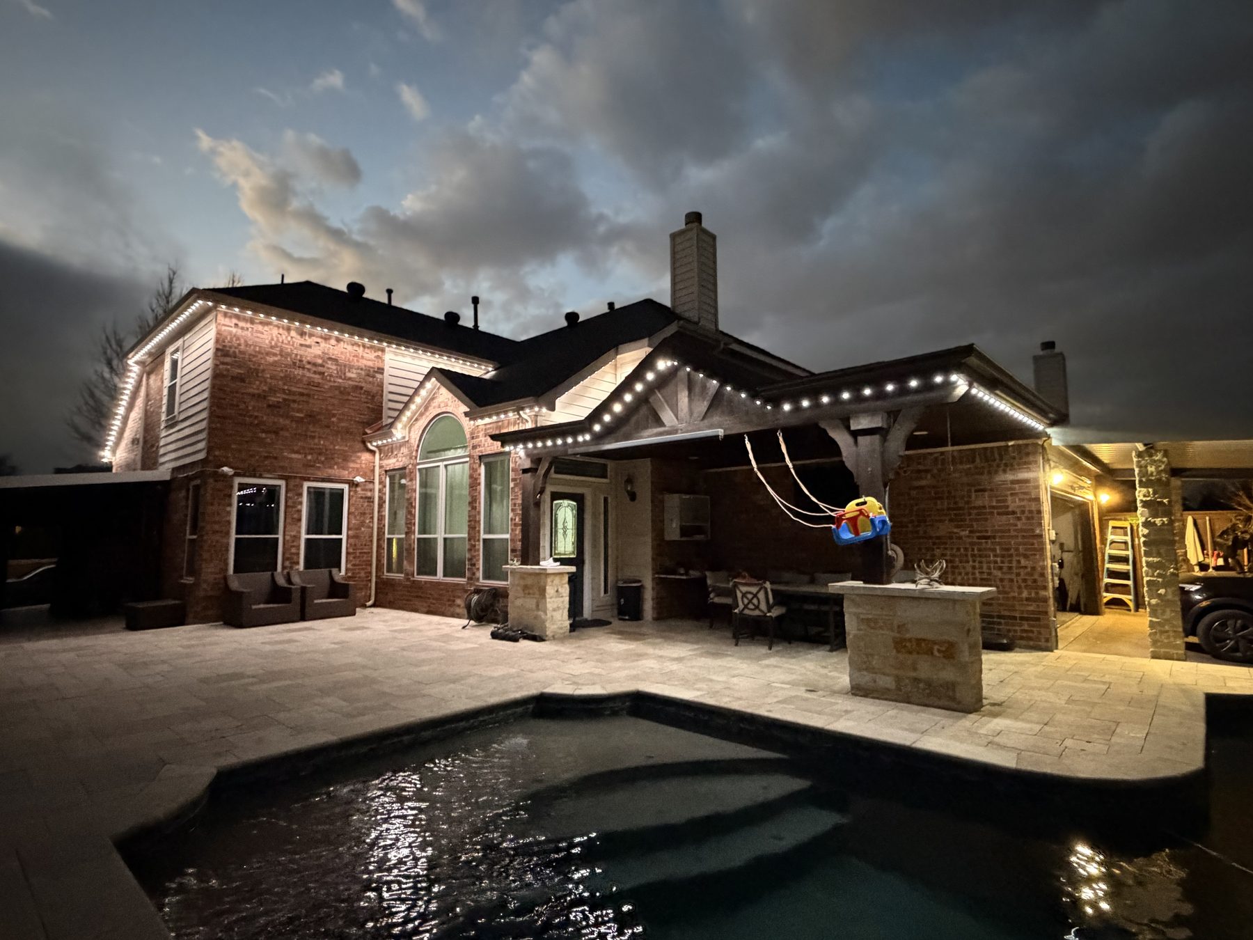 Backyard pool and covered patio lit with warm white permanent lighting